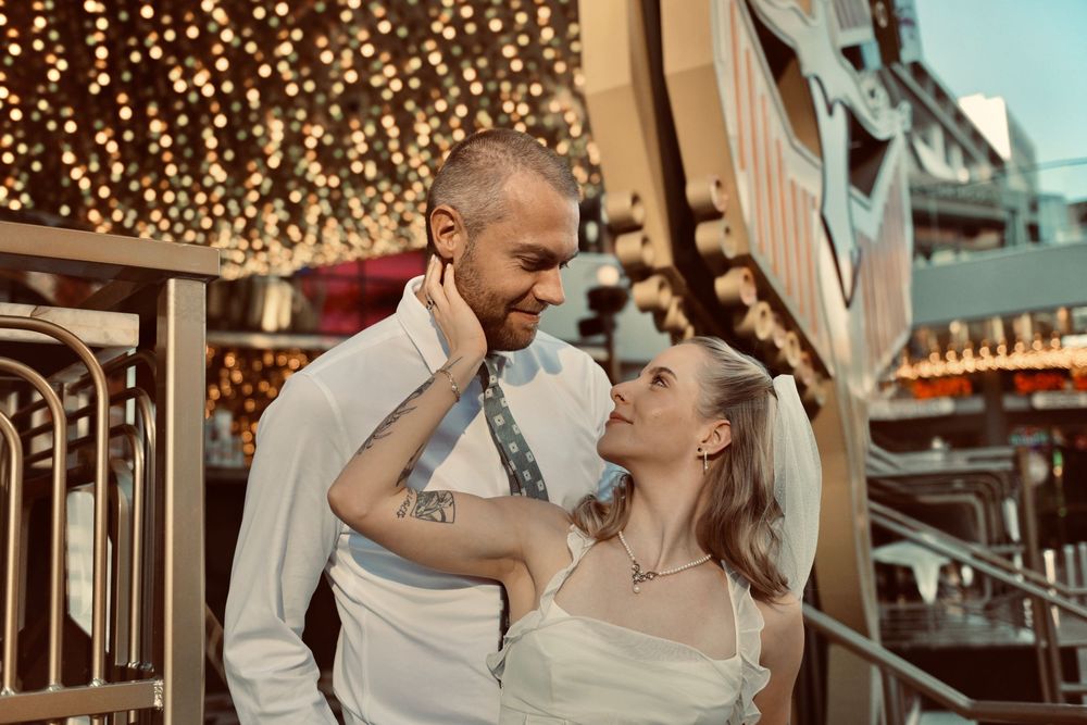 A smiling bearded man in a white shirt and tie embraces a tattooed woman in a white dress under glowing string lights.