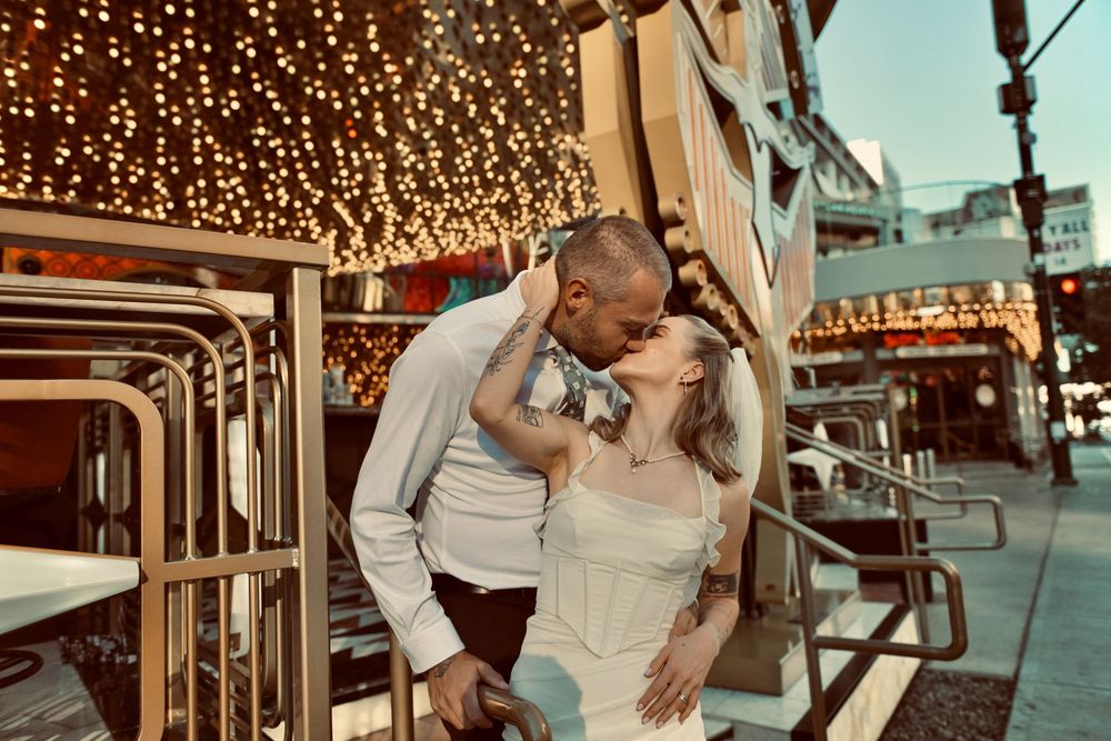 A couple in white embraces and kisses on a city sidewalk beneath twinkling string lights at dusk.
