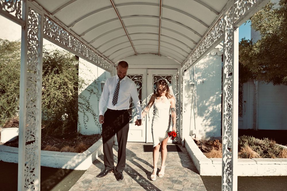 Couple in wedding attire walking hand in hand through an ornate white pergola with decorative latticework on a sunny day.