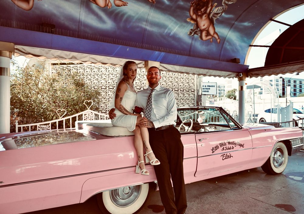 A couple poses together on the back of a pink Cadillac convertible beneath a colorful cherub-painted ceiling at a wedding chapel.