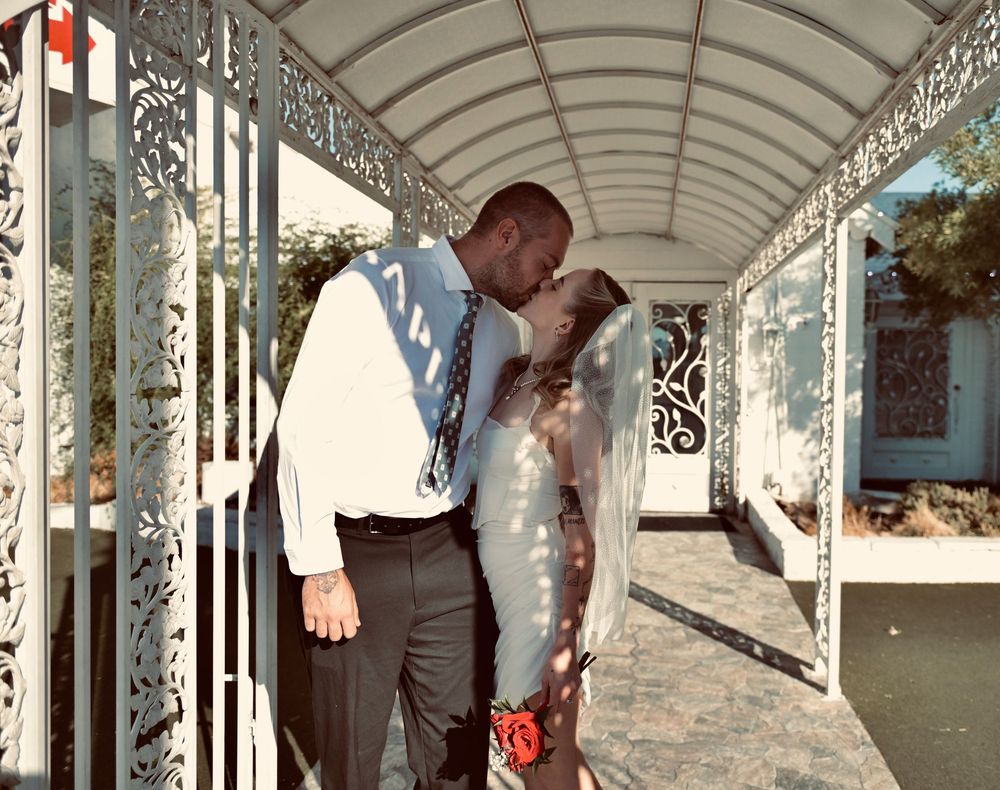 Bride in a short white dress and veil kisses her groom in a white shirt and tie beneath an ornate covered walkway.