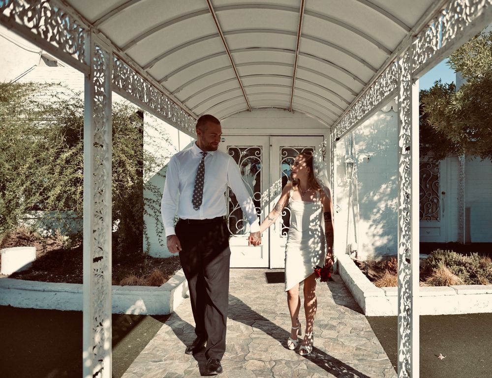 Couple holding hands walk beneath an ornate white covered walkway toward a chapel entrance, the bride in a short white dress.