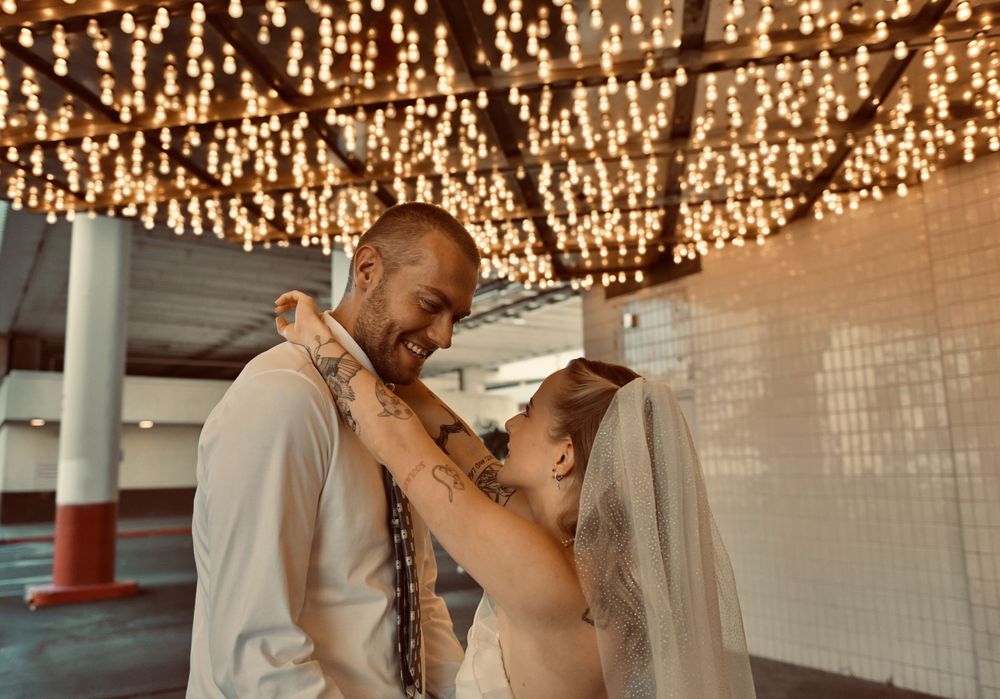 A smiling tattooed groom embraces a bride in a veil during an intimate first dance beneath a canopy of twinkling string lights.