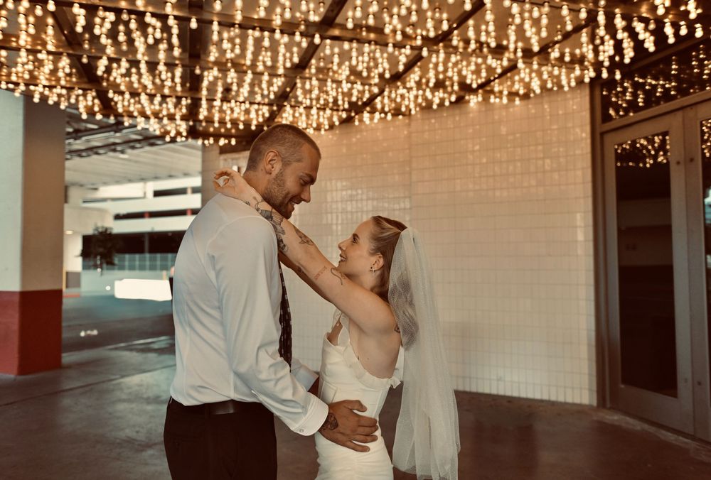 Bride in a white dress and veil embraces her groom in a white shirt and tie under a canopy of twinkling string lights.