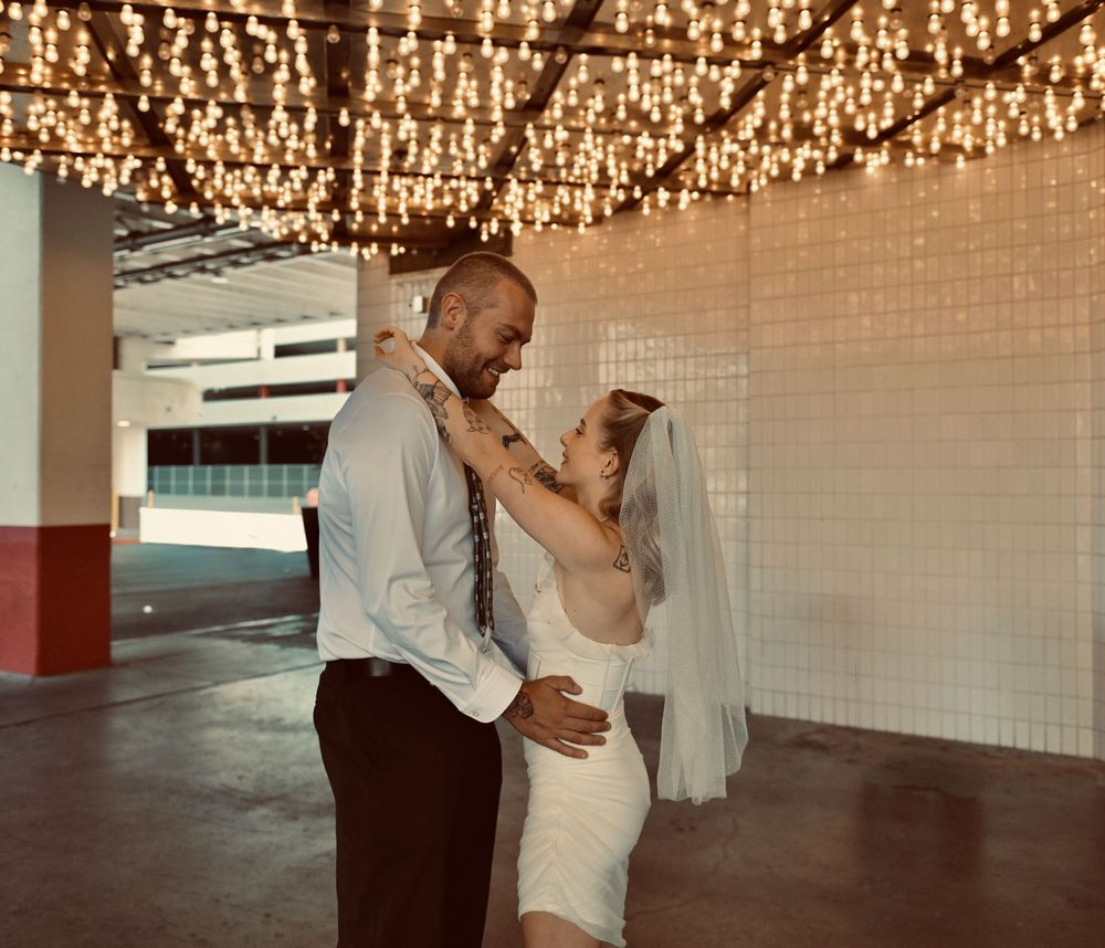 Bride in a white dress and veil dances with her groom under a canopy of twinkling string lights in an open venue.
