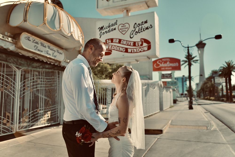 Bride in a white gown and veil embraces her groom outside a Las Vegas wedding chapel with a pink sign and the Stratosphere tower behind.