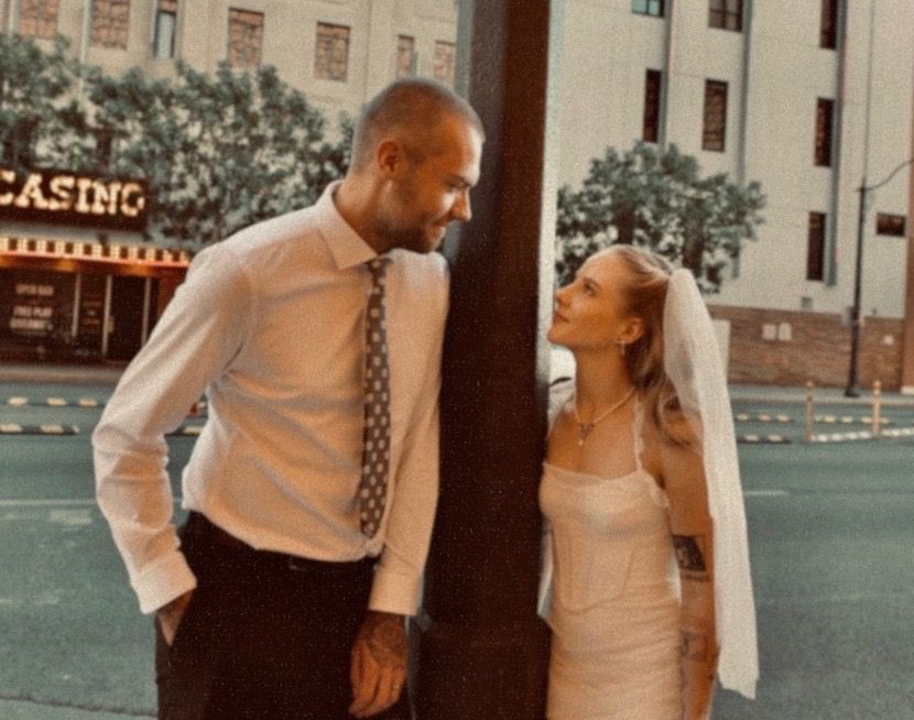 Bride in a white dress and veil smiles up at a groom in a white shirt and tie as they stand beside a pole on a city street near a casino.