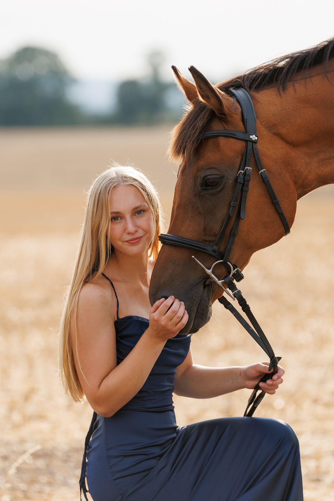 Young woman with long blonde hair in a navy satin dress kneels beside a bay horse in a bridle in a golden field at sunset.