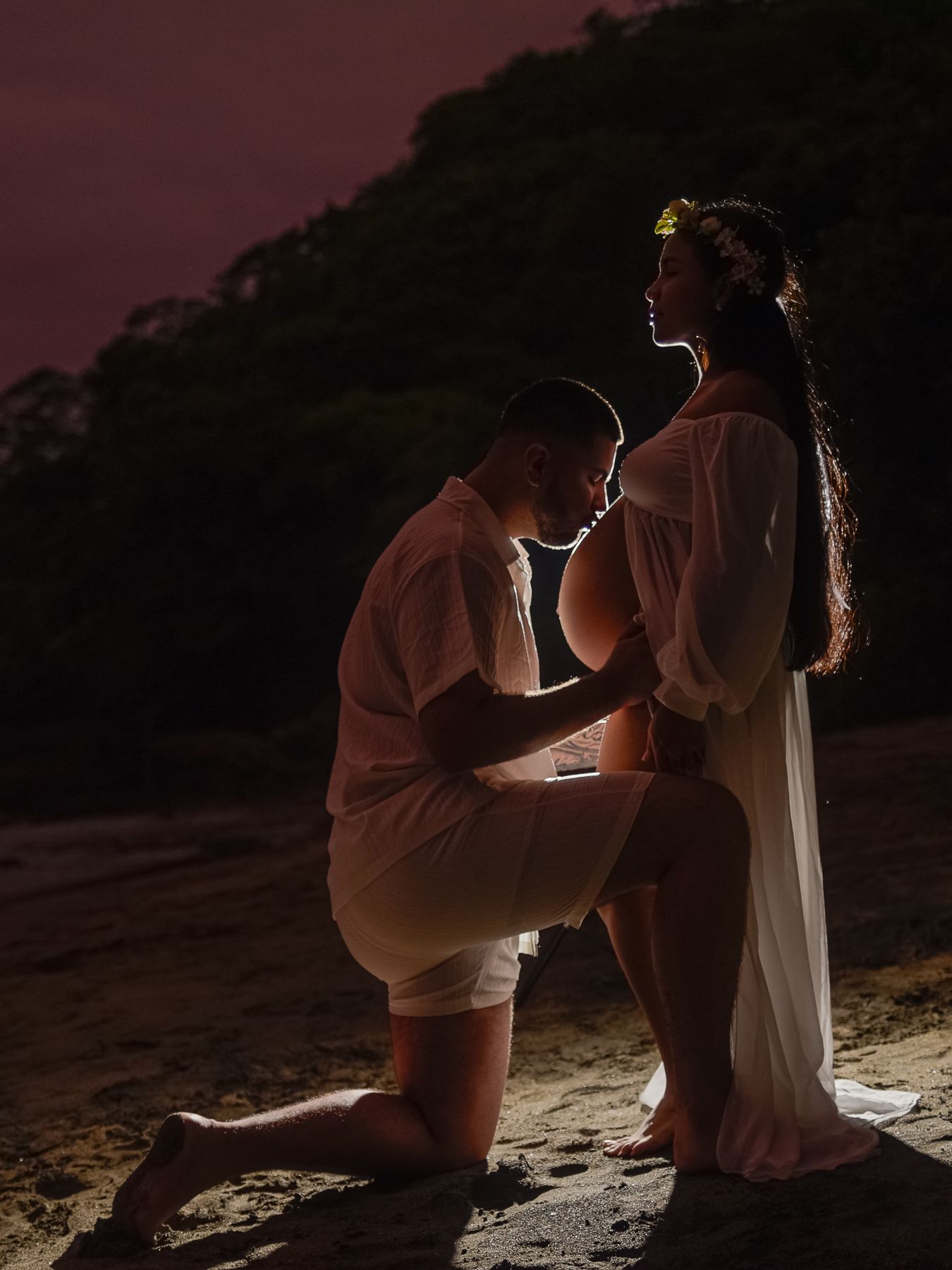 A man kneeling before a pregnant woman in a white dress on a beach at dusk, with soft backlight and lush trees behind at Waldorf Astoria