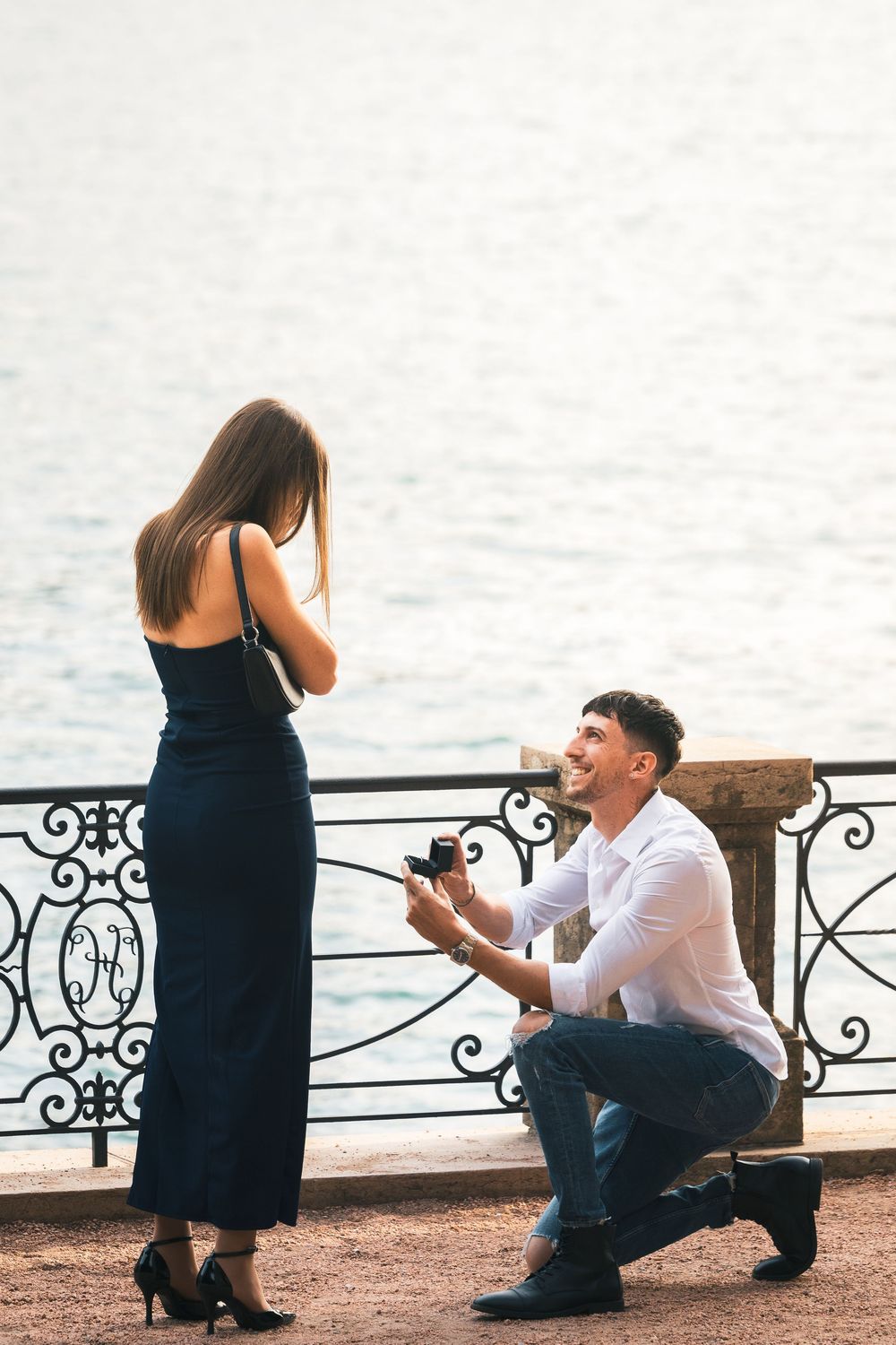 Man in white shirt kneels on one knee holding a ring box, proposing to a woman in a navy dress beside an ornate railing overlooking a lake.