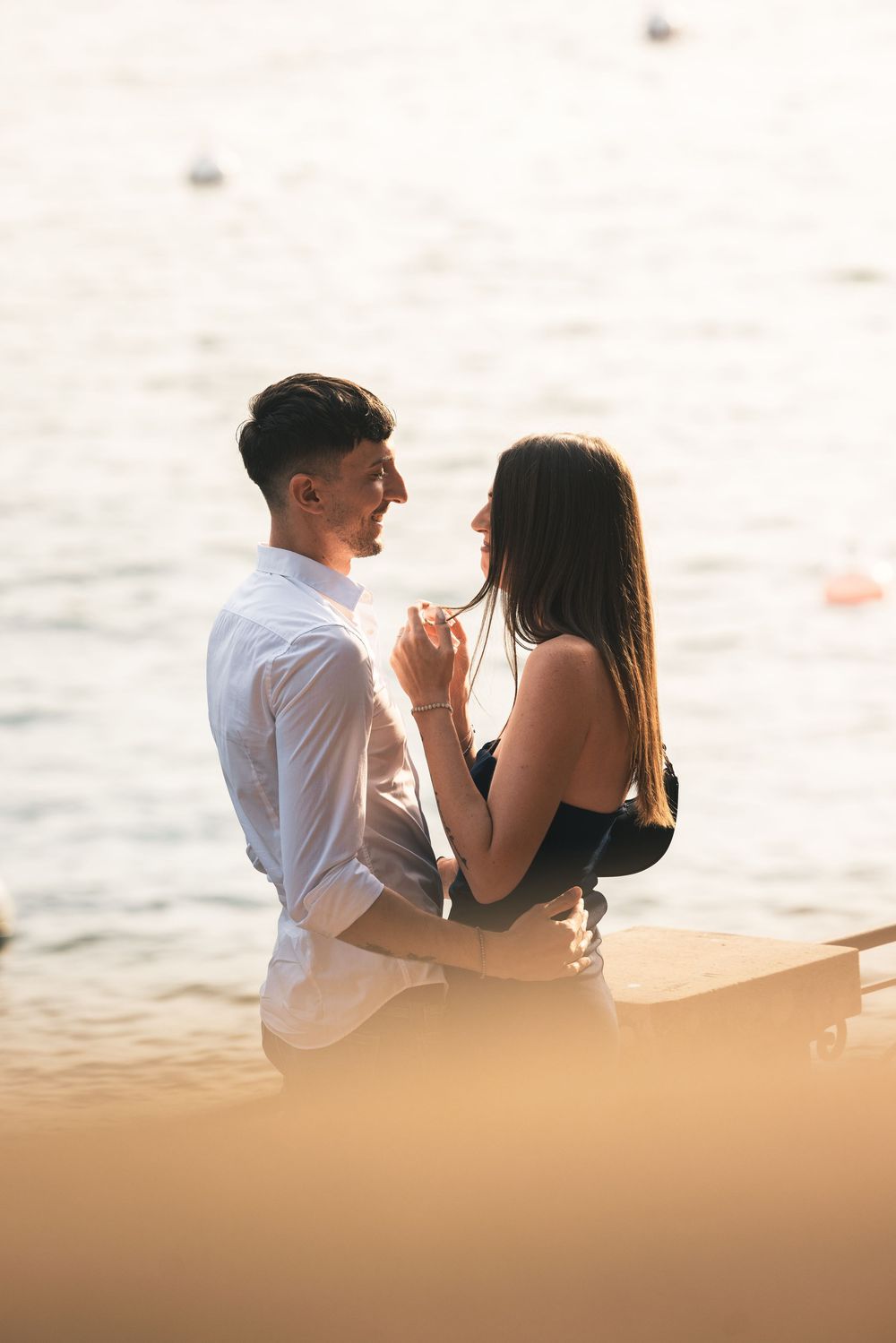 Romantic couple embracing and smiling at each other on a lakeside pier at sunset, with soft golden light glowing over the calm water.