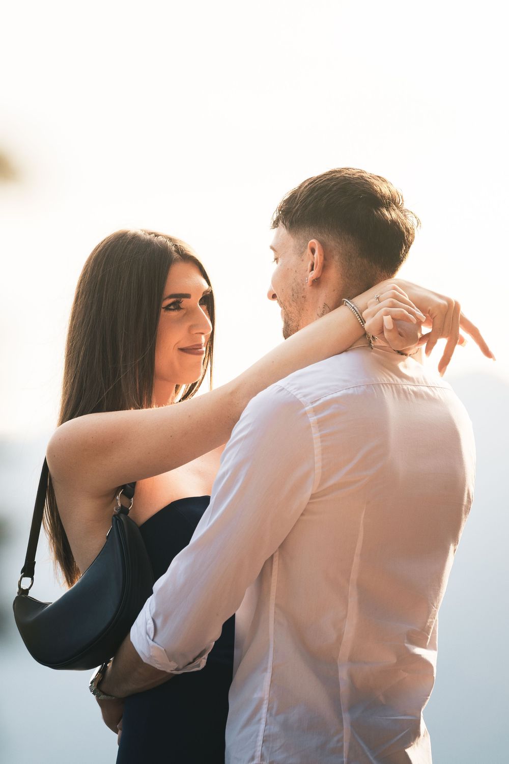 Smiling woman in a dark dress with a black shoulder bag embraces a man in a white shirt in warm, soft sunlight.