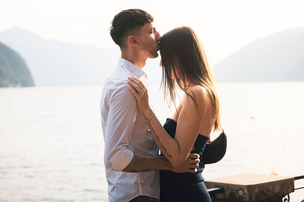 A man in a white shirt kisses the forehead of a woman in a dark dress as they embrace by a calm lake with mountains in the background.