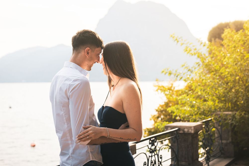 Romantic couple embracing forehead to forehead on a scenic lakeside terrace with mountains and golden sunset light in the background.