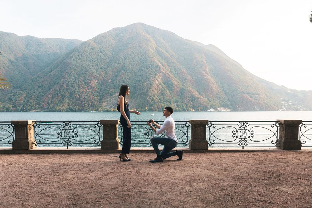Man kneeling on one knee proposing to a standing woman on a lakeside terrace with a stone balustrade and misty mountains behind.