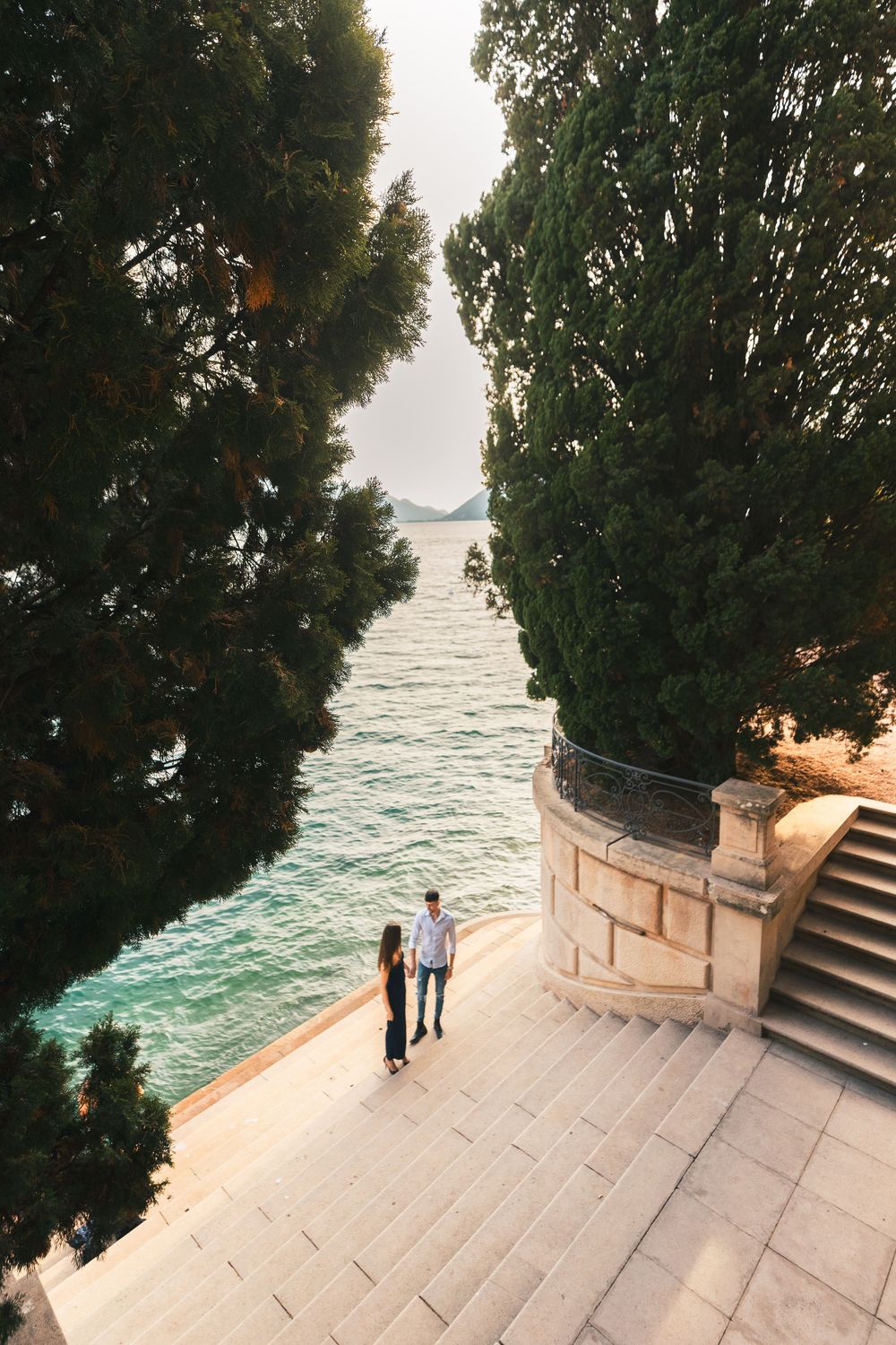 Couple standing on wide stone steps beside tall cypress trees, overlooking turquoise seaside waters at sunset.