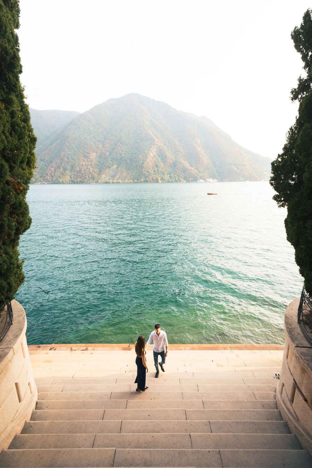 Couple walking down wide stone steps toward a turquoise lake with forested mountains and cypress trees in the background.