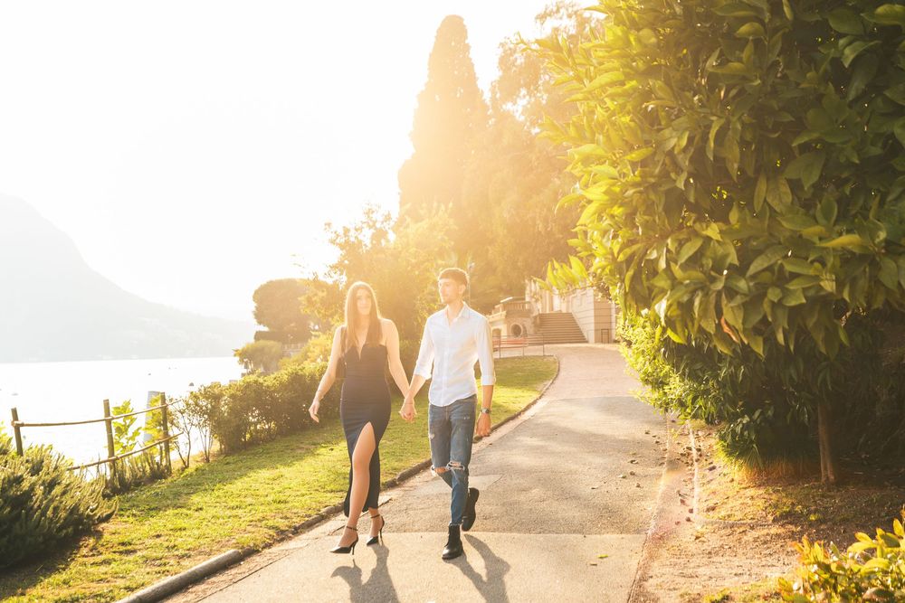 Couple holding hands and walking along a sunlit lakeside path surrounded by lush green trees during a romantic golden hour stroll.