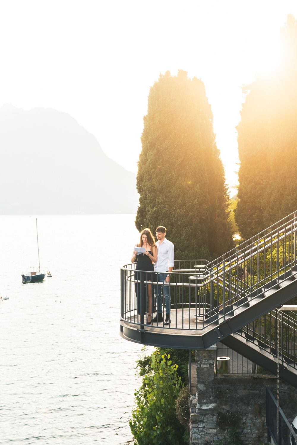 Couple stands on a metal balcony with staircase overlooking a calm lake and sailboat at golden sunset, with tall cypress trees nearby.