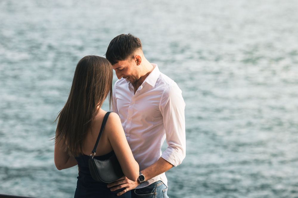 A couple embraces closely by the water, the man in a white shirt smiling down at the woman in a dark top in a romantic moment.