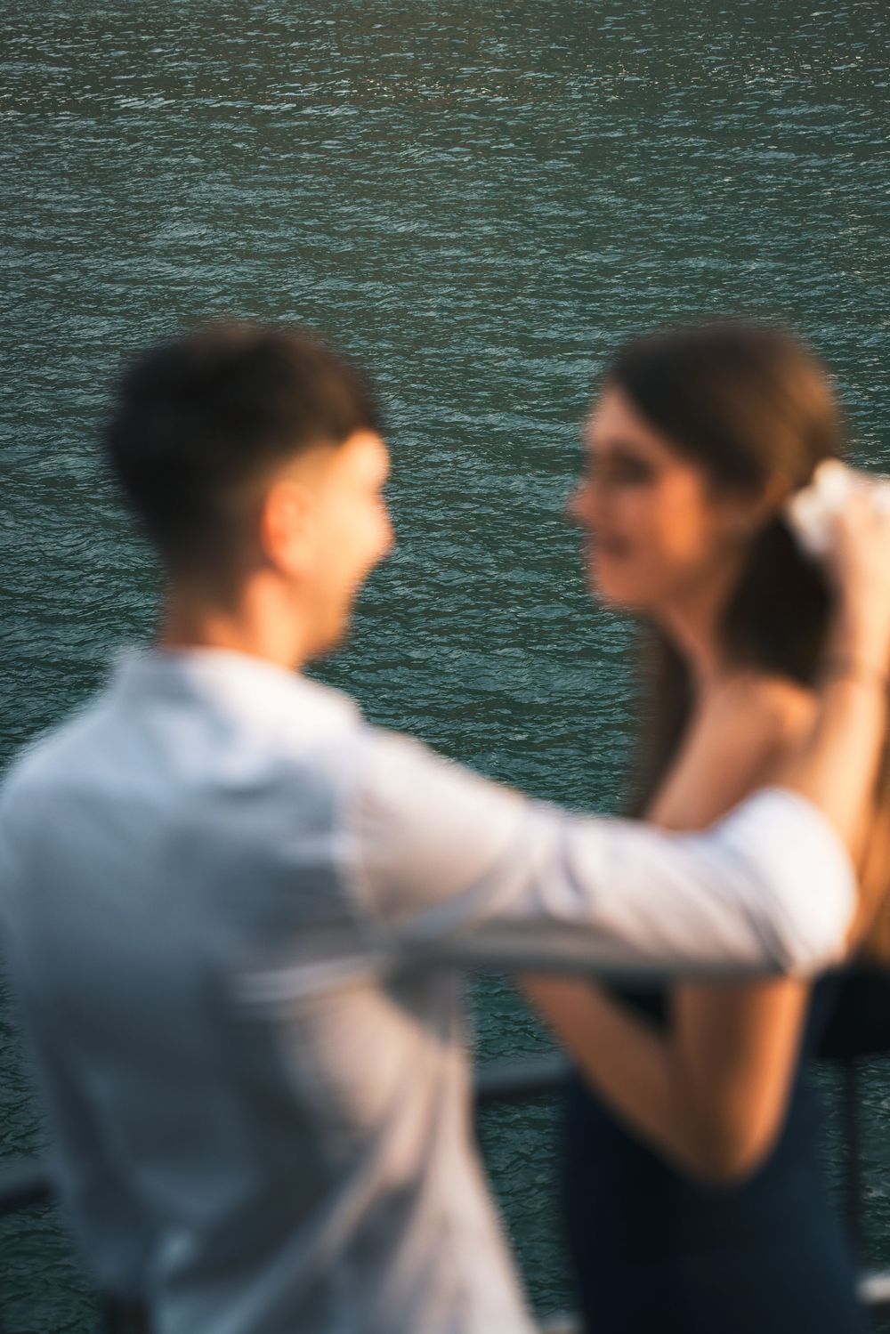 Blurred romantic couple smiling at each other on a waterfront terrace, the woman in a navy dress reaching up to her hair, lake behind them.