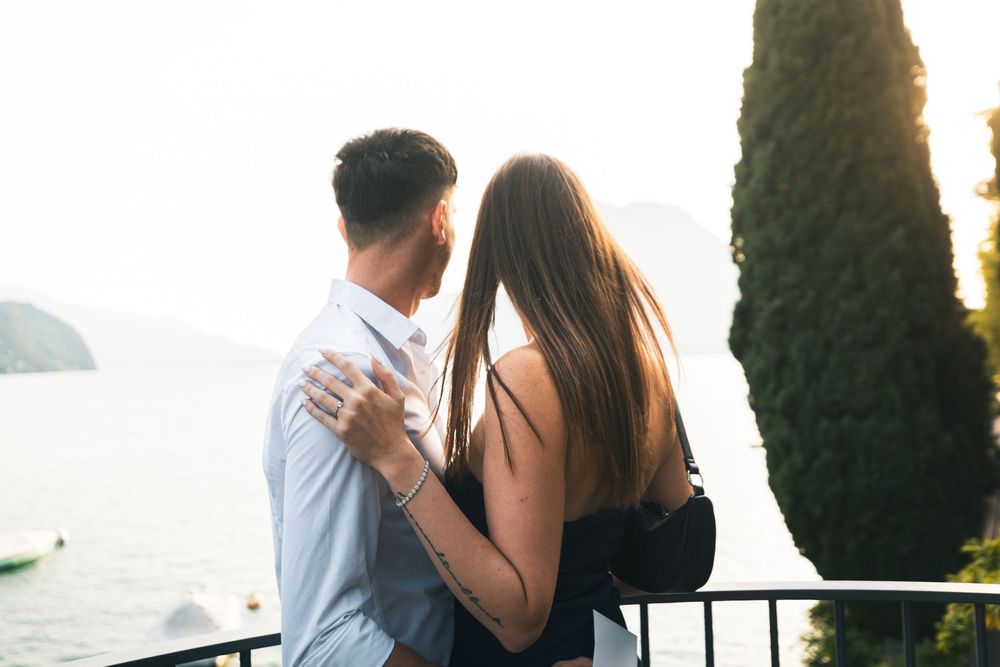 Couple embracing from behind on a balcony overlooking a calm lake, with distant mountains and a tall cypress tree in the soft evening light.