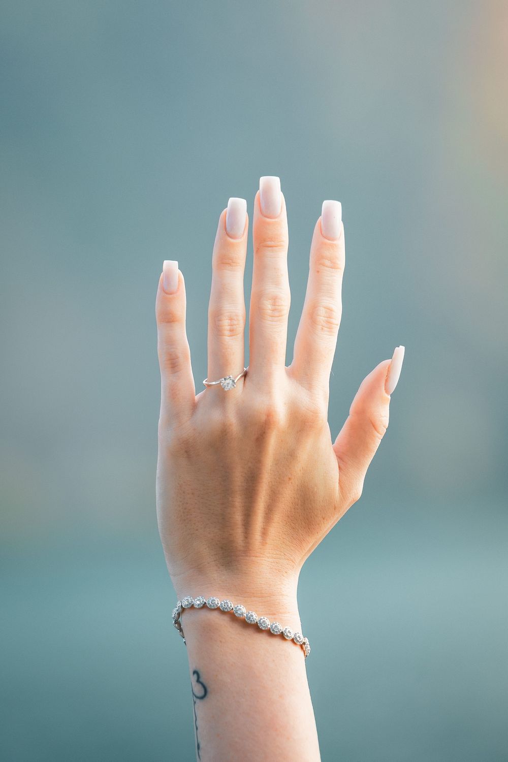 Raised hand with white manicured nails, a diamond engagement ring and a sparkling tennis bracelet against a soft teal blurred background.