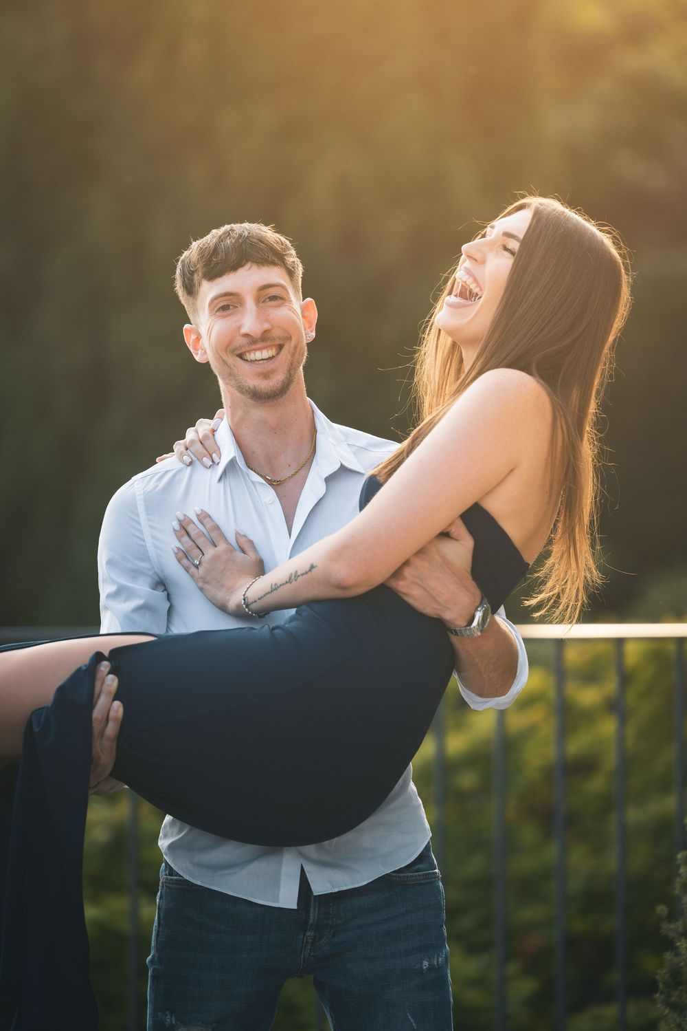 Smiling man in a light blue shirt lifts a laughing woman in a black dress outdoors during golden hour sunlight.