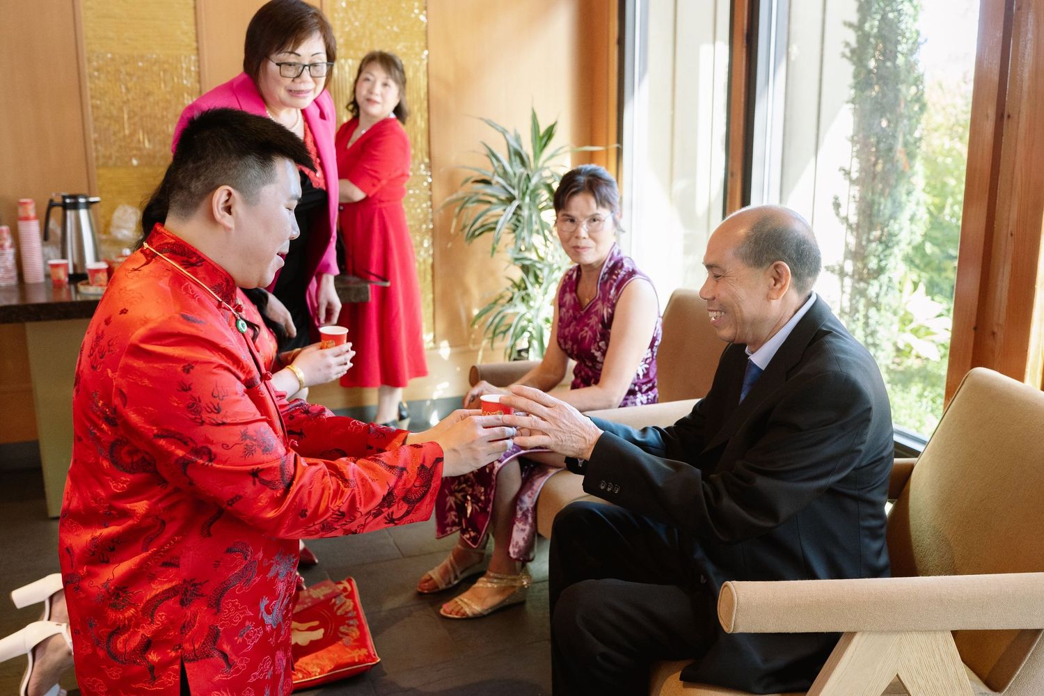 A groom in a red traditional jacket offers tea to a seated elder indoors, with family members seated nearby.