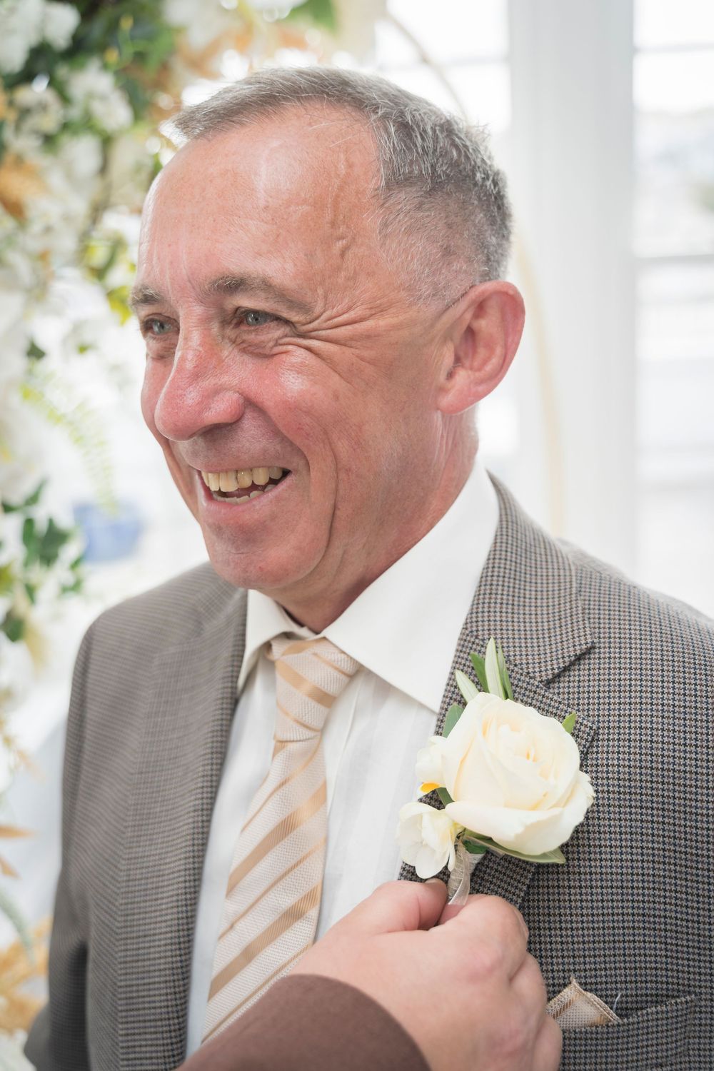 Smiling man in a gray tweed jacket and striped tie has a white rose boutonniere pinned to his lapel at a wedding.