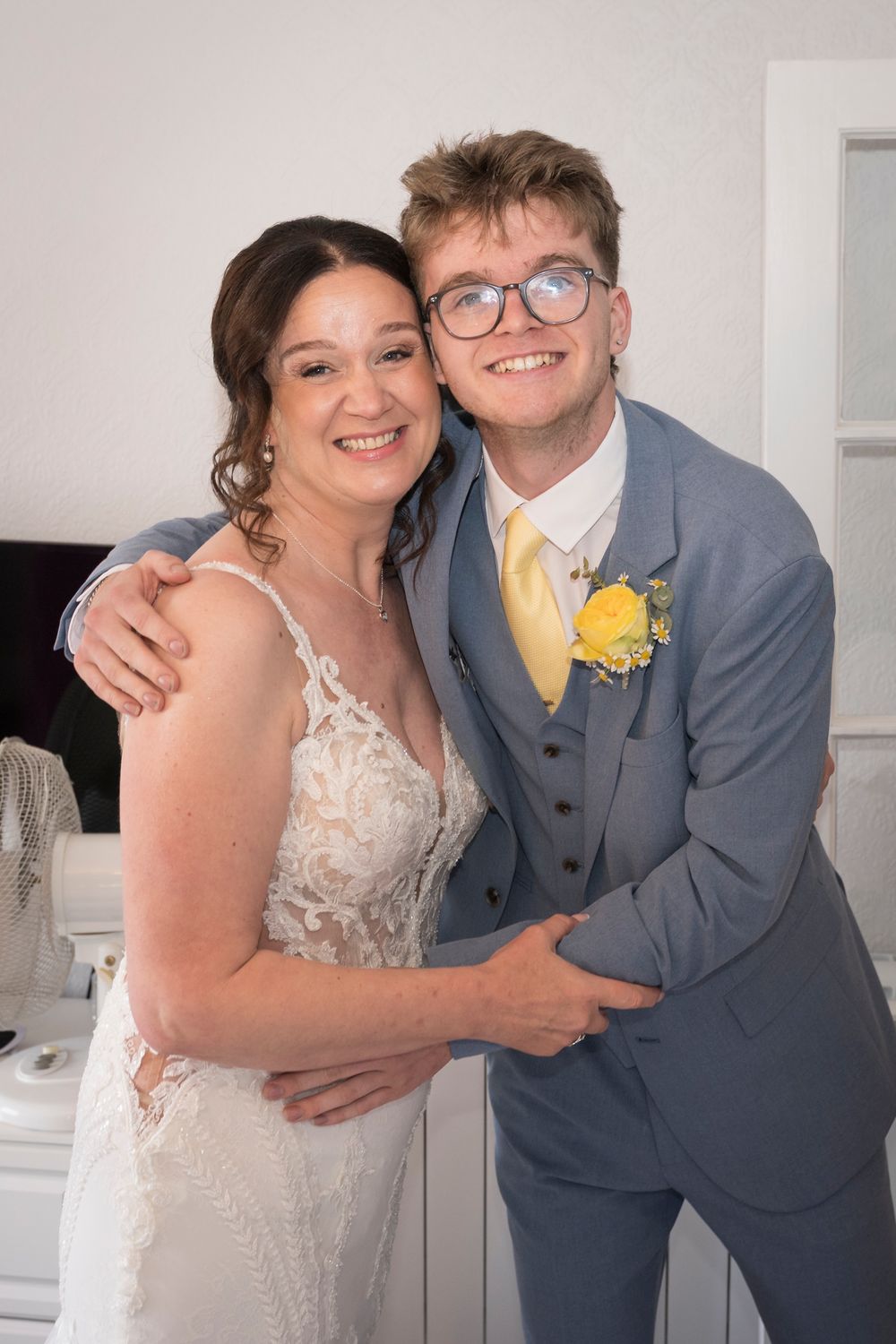 Smiling woman in a lace wedding dress embraces a young man in glasses wearing a gray suit with a yellow tie and boutonniere.