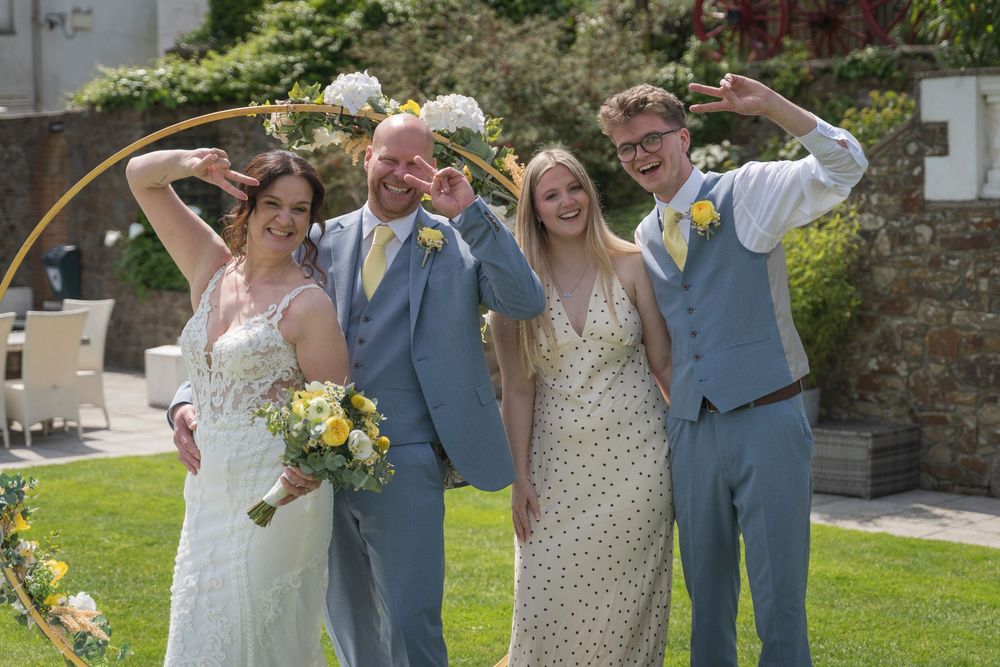 Smiling bride in a white lace gown and groom in a light blue suit pose with two guests holding a bouquet at an outdoor garden wedding.