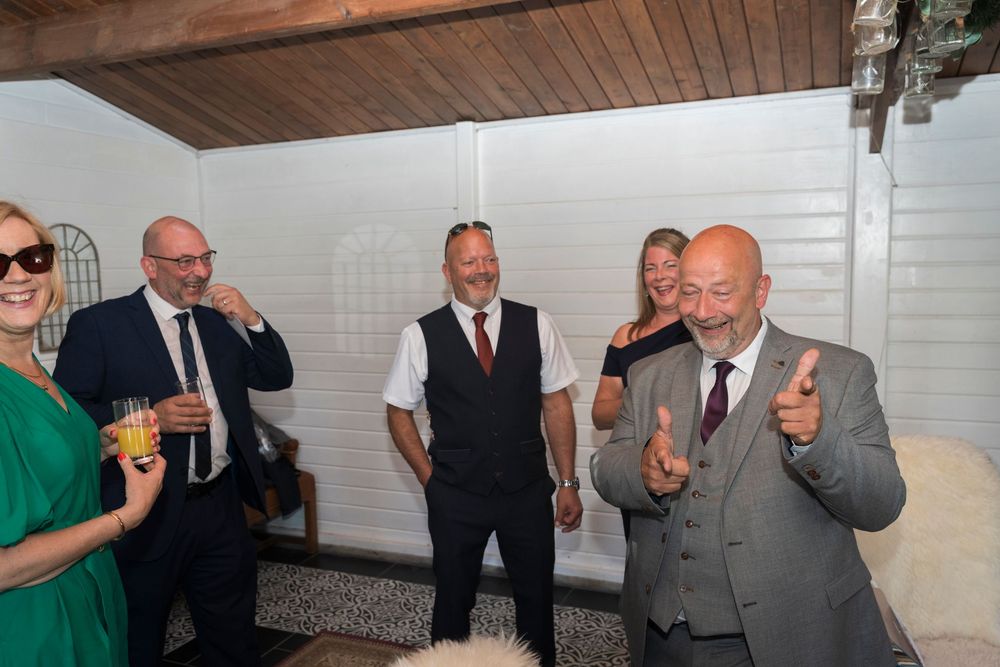 A bald man in a gray suit gives two thumbs up at a lively indoor party as smiling guests in formal attire mingle behind him.