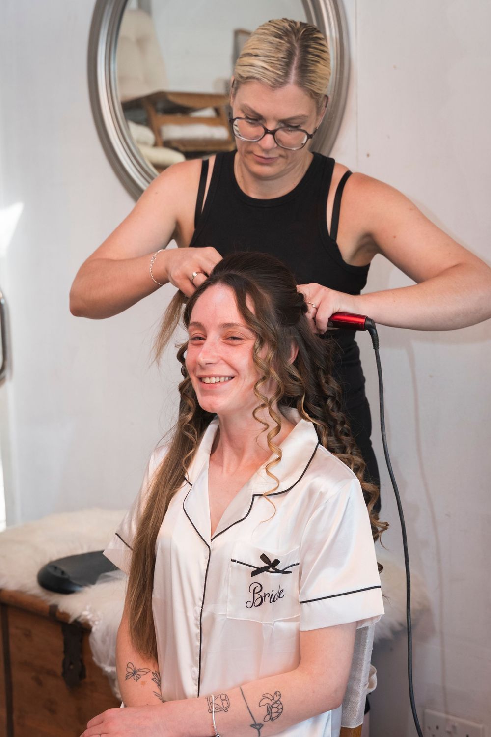 Smiling bride in a white satin "Bride" robe sits while a hairstylist curls her long hair with a curling iron on her wedding morning.