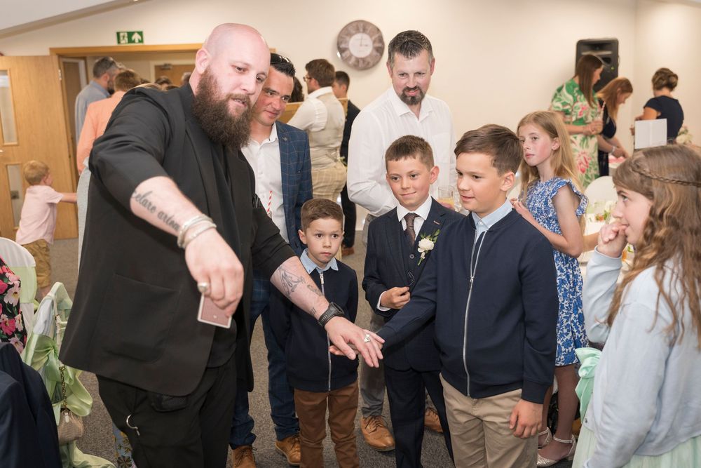 A bearded man in black performs a card trick for a group of smiling children in dress clothes at an indoor family event.