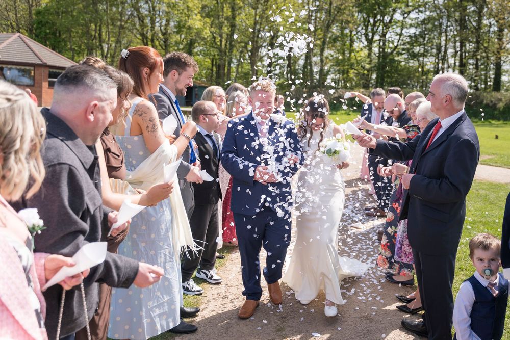 Bride and groom walk through a cheering crowd of guests throwing confetti outdoors at a sunny wedding ceremony.