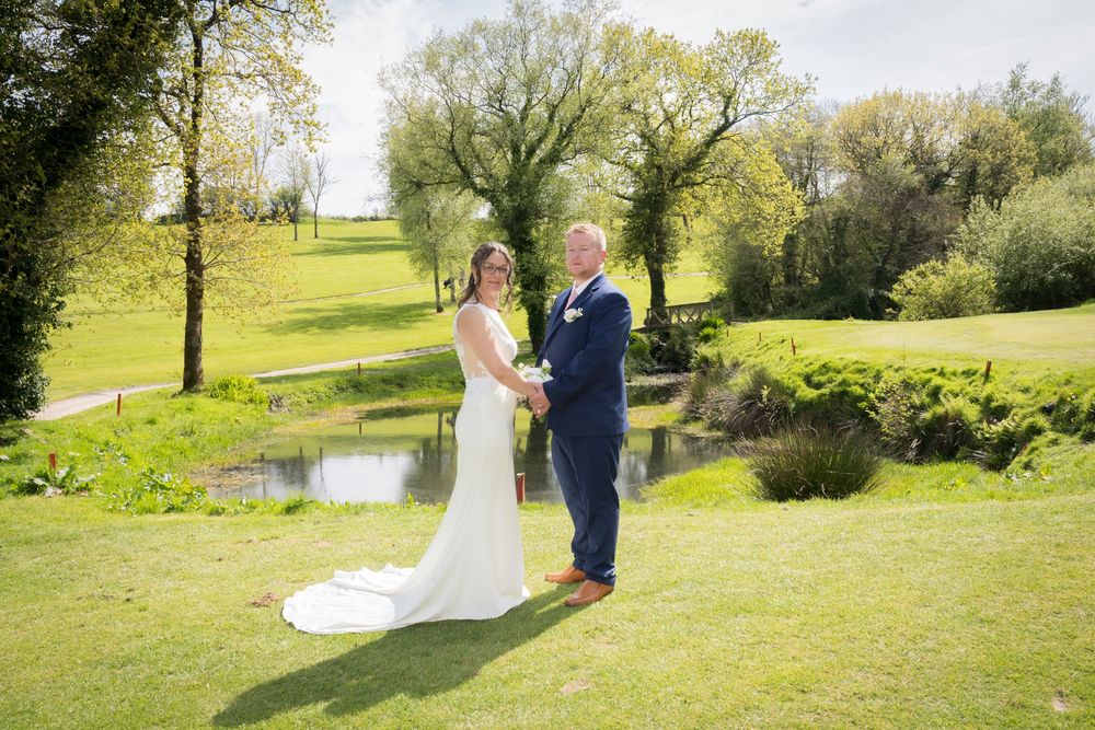Bride in a white gown and groom in a navy suit hold hands on a sunny green lawn beside a small pond at a scenic wedding venue.