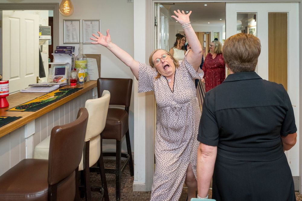 A joyful woman in a patterned dress raises her arms in celebration inside a bright café while another woman in black watches.