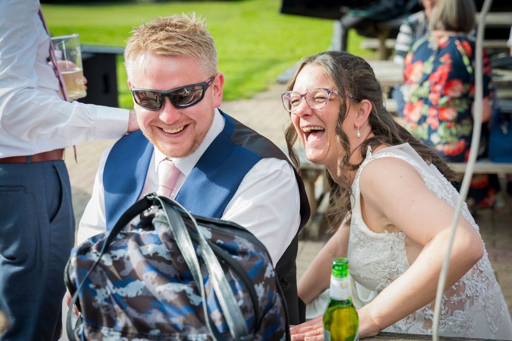 Smiling man in sunglasses and blue vest laughs with a woman in glasses at an outdoor wedding reception table with a green bottle.