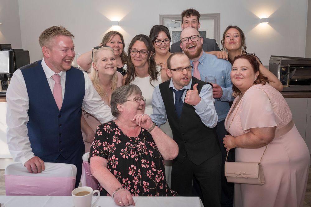 Smiling group of family and friends in formal attire posing closely for a joyful celebration photo, with one man giving a thumbs up.