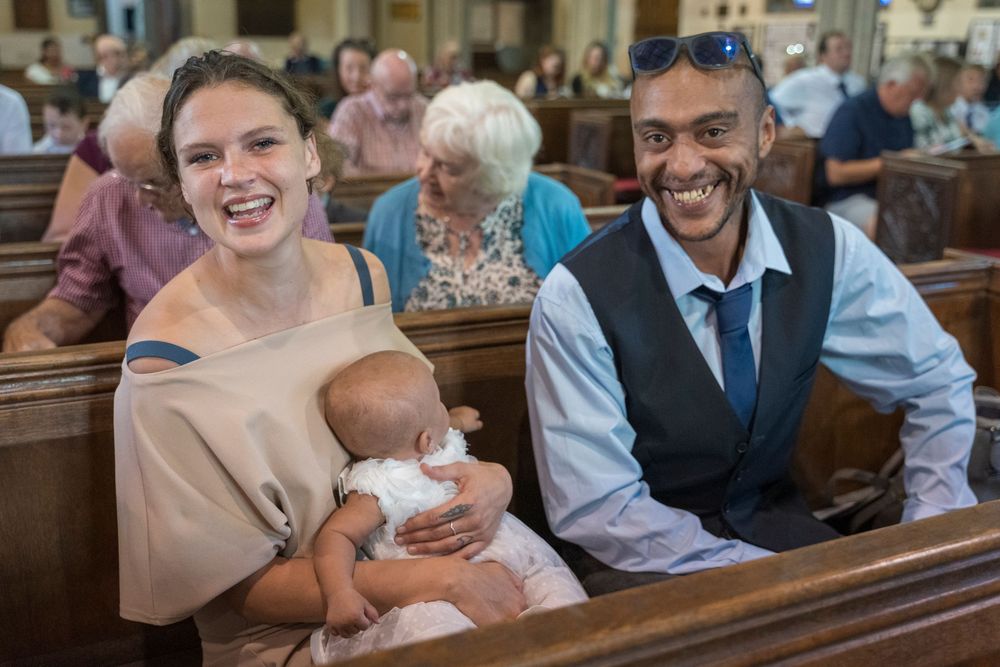 Smiling woman holding a baby sits beside a smiling man with sunglasses on his head in a church pew, with other worshippers behind them.