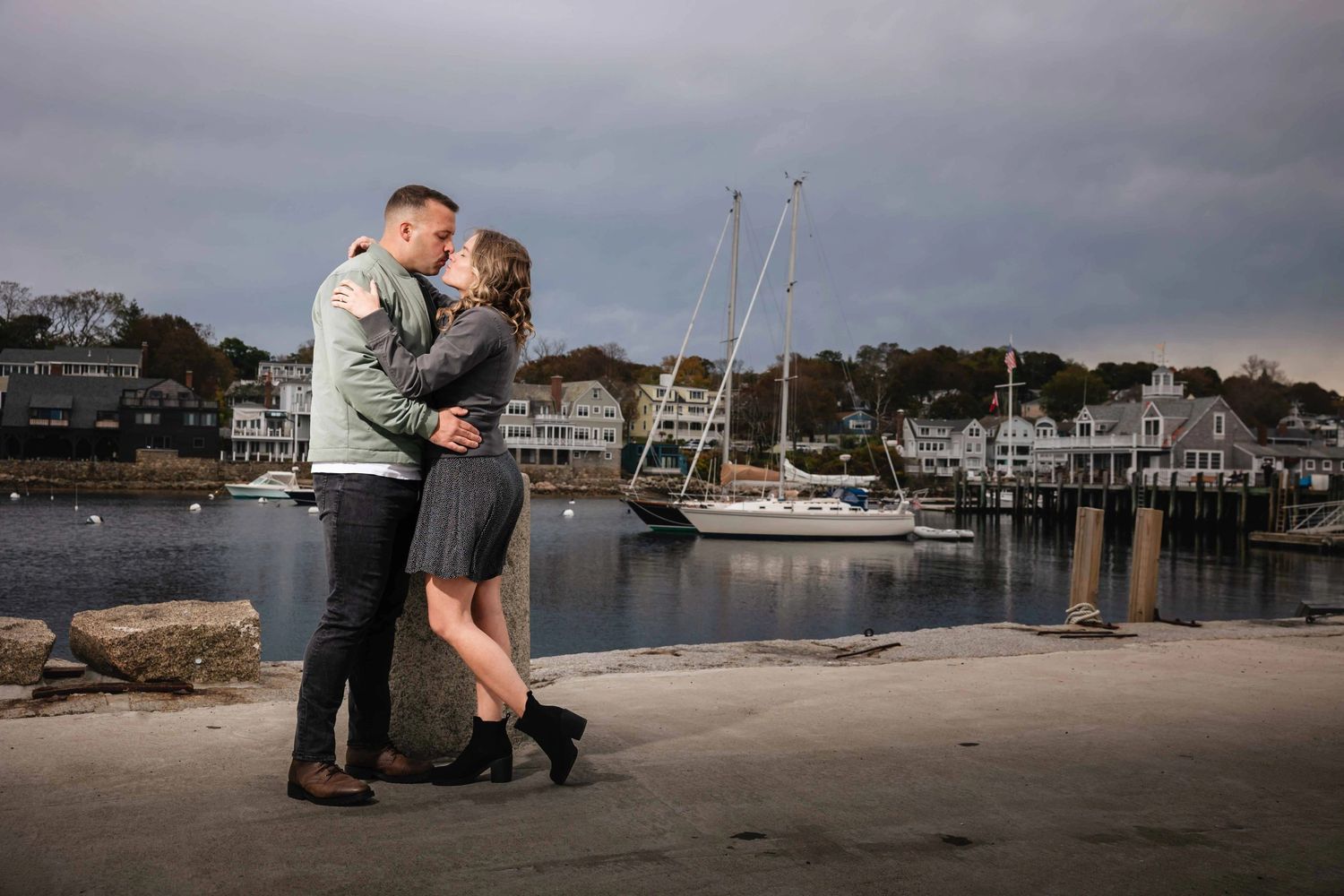 Couple embracing and about to kiss on a sandy beach with sailboats moored behind them. Engagement at Rockport Harbor, Rockport, MA