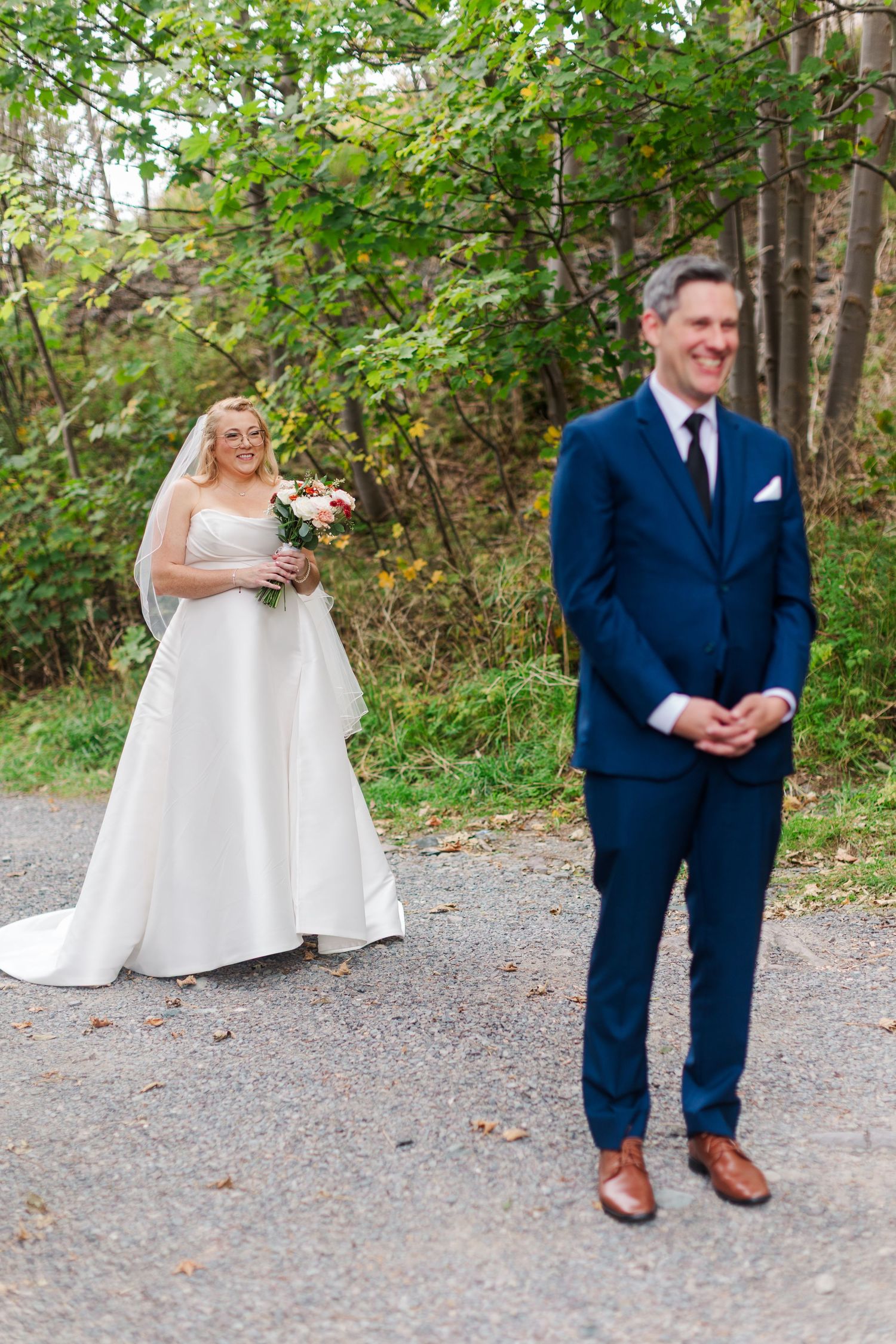 Bride in a white gown holding a bouquet watches smiling as the groom in a navy suit stands facing away on a tree-lined path.