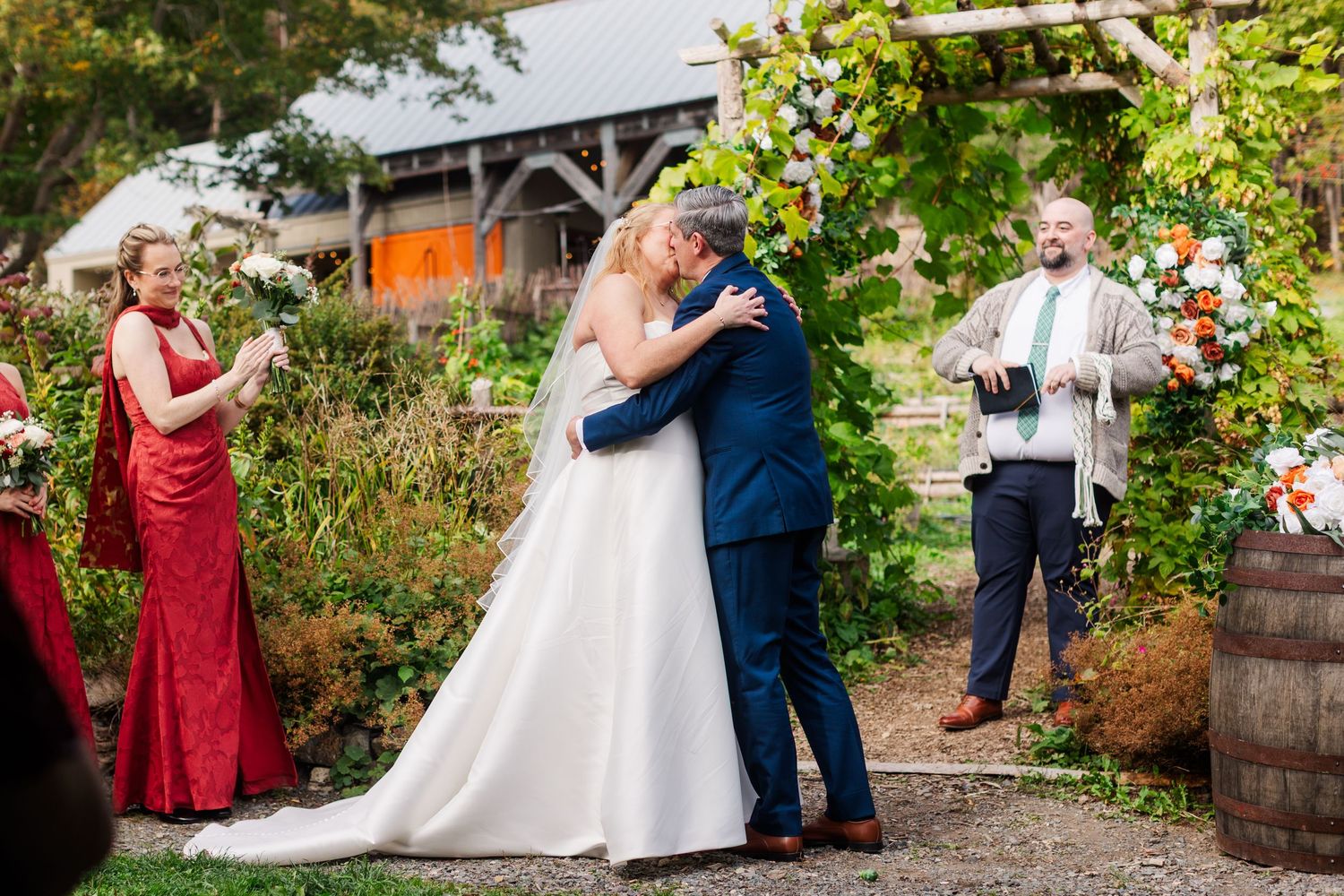 Bride in a white gown and groom in a navy suit kiss under a flower-covered arch as a bridesmaid in red and an officiant look on.