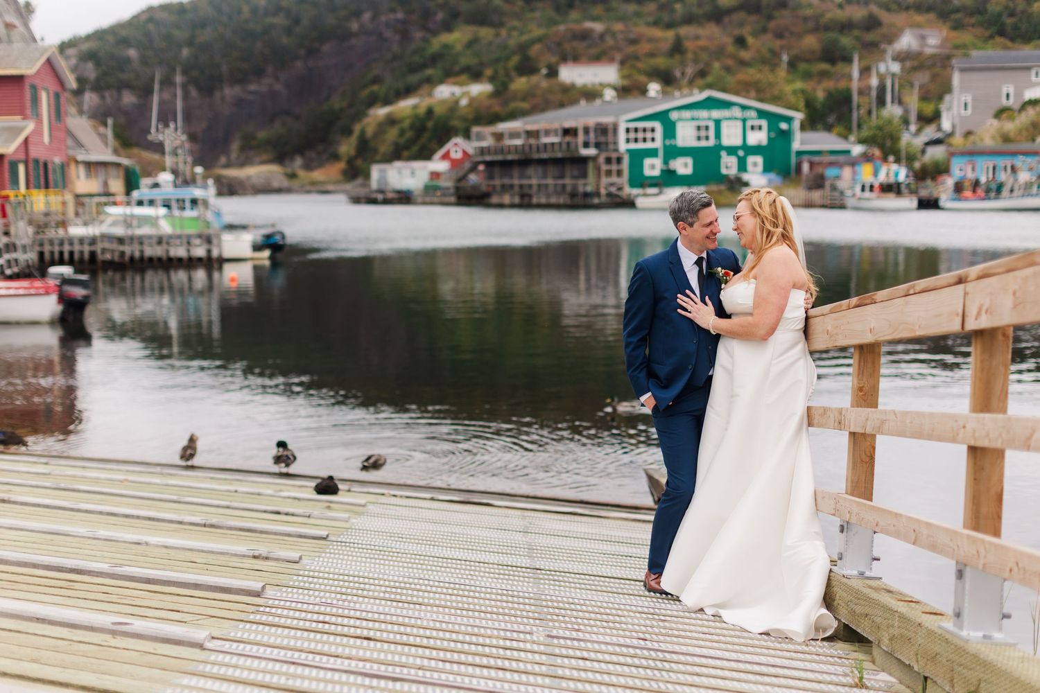 Bride in a white gown and groom in a navy suit embrace on a wooden dock overlooking a calm harbor with colorful waterfront houses.
