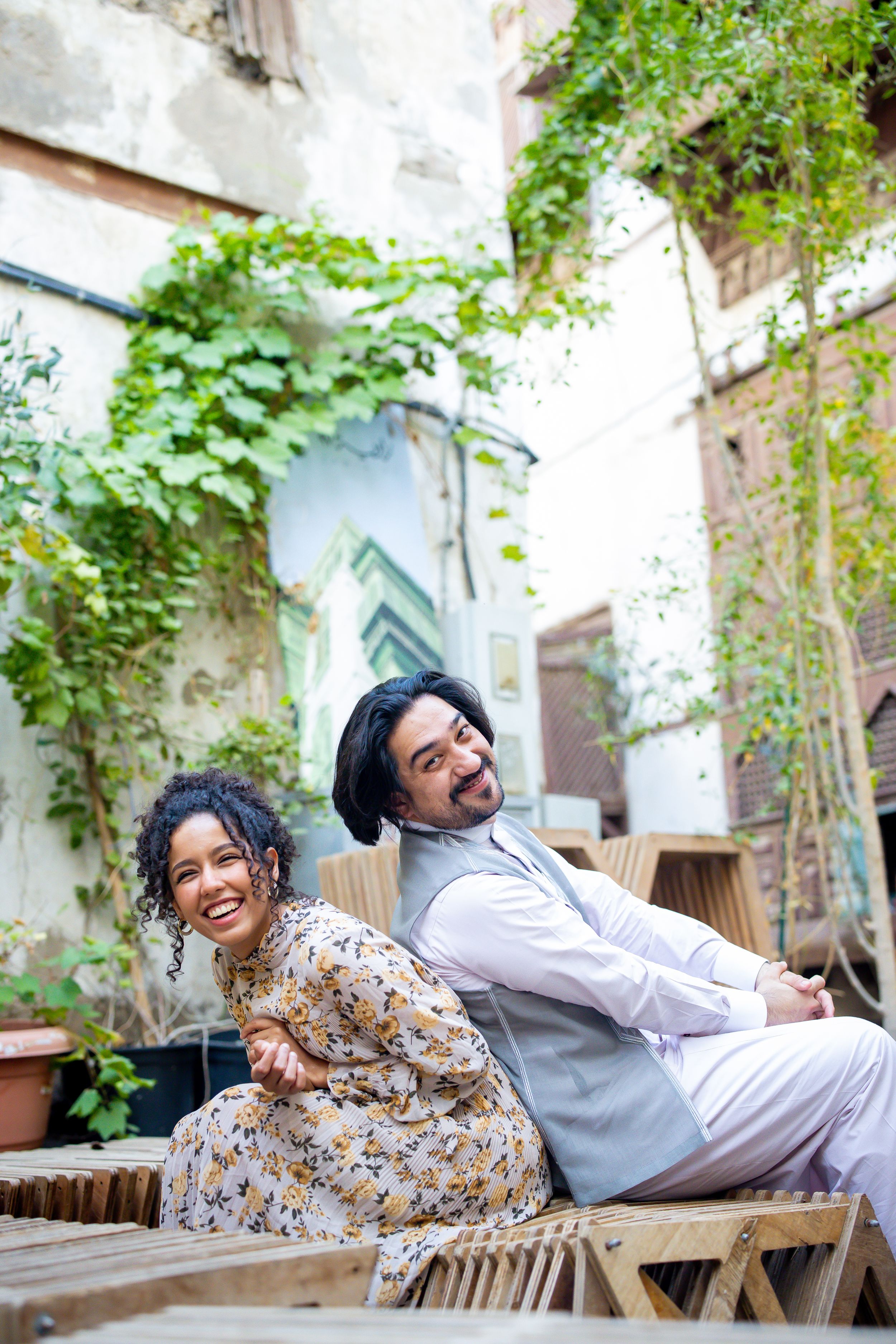 Smiling couple sitting back-to-back on wooden furniture in a charming courtyard with green ivy-covered walls.