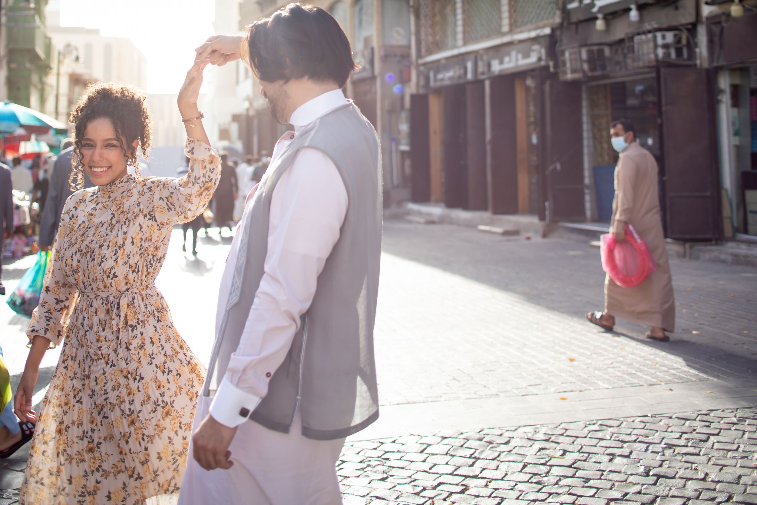 Couple dancing together on a sunny urban street, woman in floral dress, man in gray vest, holding hands outdoors.