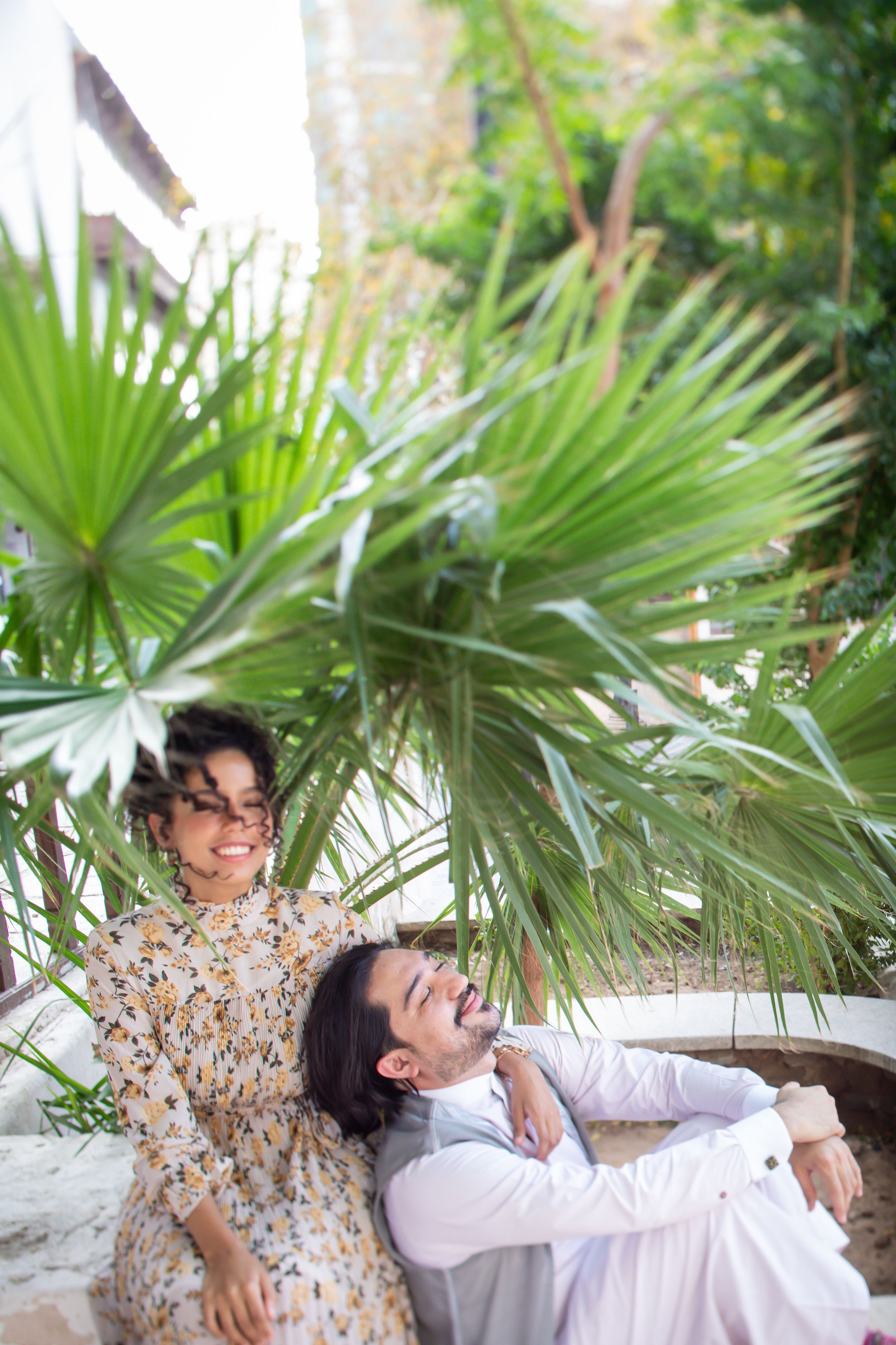 A happy couple relaxing together among lush tropical palm plants, woman in floral dress, man in white outfit.