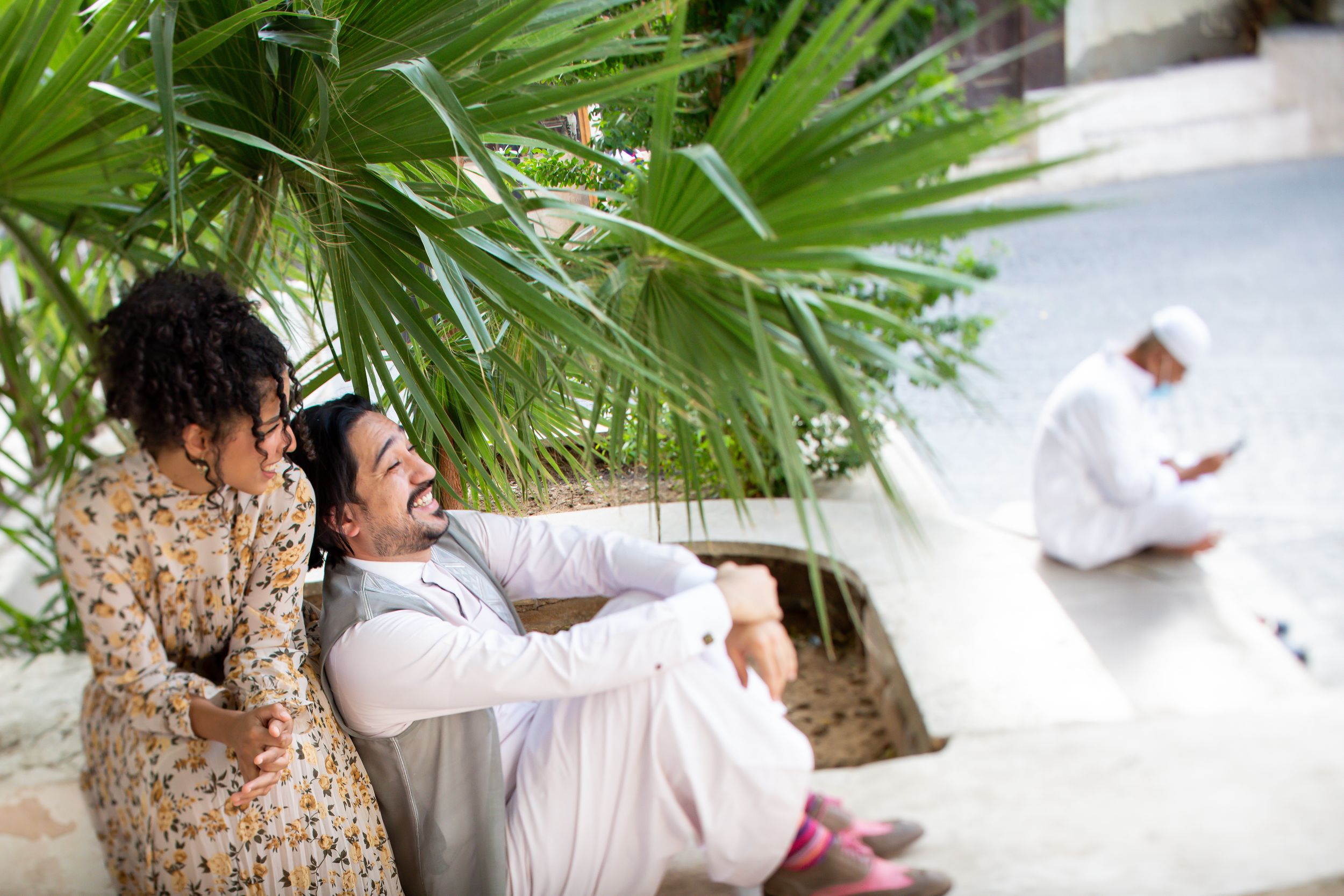 A man and woman in traditional Middle Eastern attire sit together under palm trees, sharing a joyful moment outdoors.