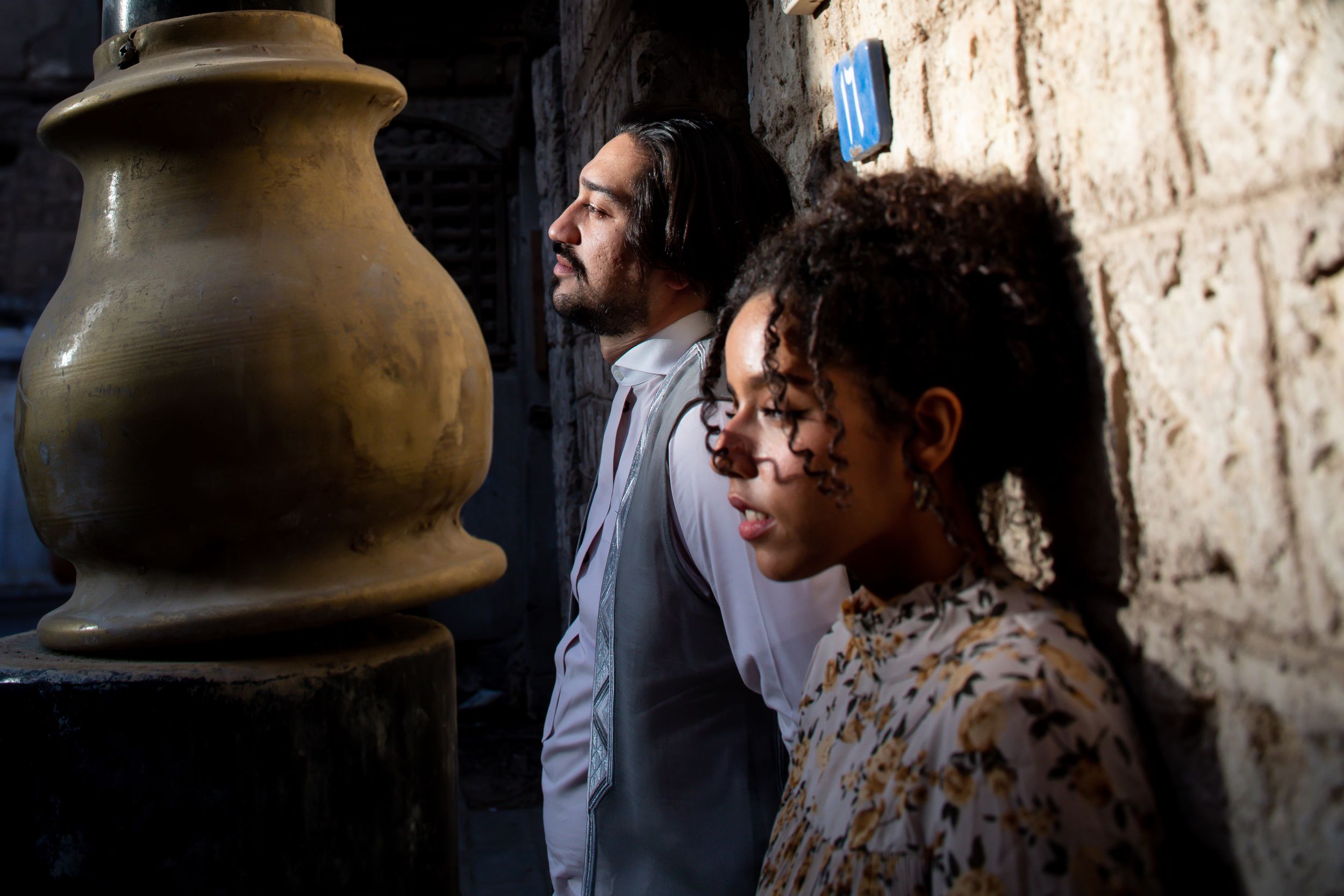 Two people with dirty faces lean against a dark stone wall near a large clay pot in a dramatic scene.